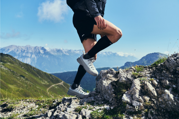 A close-up lifestyle shot of a runner's legs and feet mid-step on a rocky mountain ridge, wearing black athletic shorts, black compression socks, and white trail running shoes, with a scenic mountain landscape under a blue sky in the background.