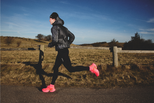 A person running outdoors on a paved path wearing a black hooded jacket, black leggings, and bright pink running shoes, with a grassy field, fence, and blue sky in the background.