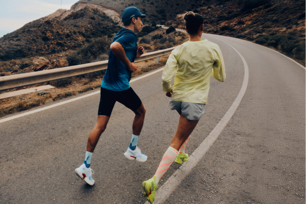 Two runners jogging on a winding mountain road, shown from behind. One runner wears a blue long-sleeve top, black shorts, white socks, and white running shoes, while the other wears a pale yellow long-sleeve top, gray shorts, and bright yellow running shoes.