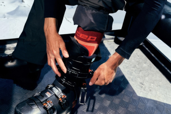A close-up of a person adjusting the strap of a black ski boot while sitting on a vehicle or platform, with snow visible in the background. The person is wearing a red and black ski sock with the brand name 'SAP' visible.