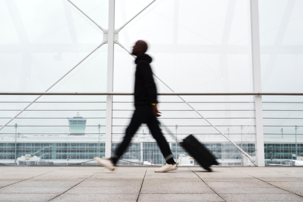 A person in a black outfit walks across a tiled floor while pulling a black rolling suitcase, photographed in front of a large glass and metal structure at an airport.